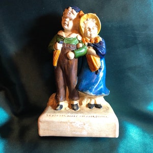 May include: A ceramic figurine of a boy and girl holding hands. The boy is wearing a green shirt, brown pants, and a blue hat. The girl is wearing a blue dress, yellow hat, and a yellow scarf. The base of the figurine is inscribed with "Mr. and Mrs. Harry Walters, Junior."