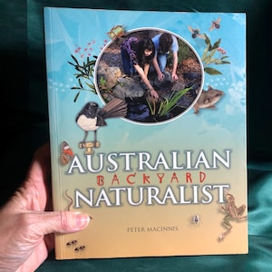 May include: A book cover with a blue and orange background. The title is "Australian Backyard Naturalist" by Peter Macinnis. The cover features illustrations of various Australian wildlife, including a bird, a frog, a spider, and a snake.