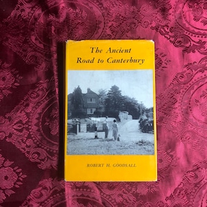 May include: A yellow book cover with the title "The Ancient Road to Canterbury" and the author's name "Robert H. Goodsall". The cover features a black and white photograph of a street scene with a house and two people walking.