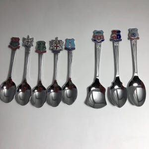 May include: A collection of eight silver-toned metal souvenir spoons arranged in two rows. Each spoon features a unique decorative emblem on the handle, displaying various colorful crests, shields, and text labels representing different locations.