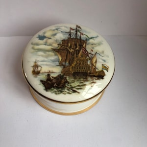 May include: A white ceramic trinket box with a gold trim. The lid features a hand-painted image of a ship with sails and flags, sailing on a blue sea with white clouds.