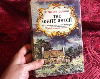 May include: A vintage paperback book cover with a green and blue illustration of a cottage with a thatched roof. The title of the book is "The White Witch" by Elizabeth Goudge. The book is a new historical novel set in the adventurous days of Charles I by the author of "Green Dolphin Country."