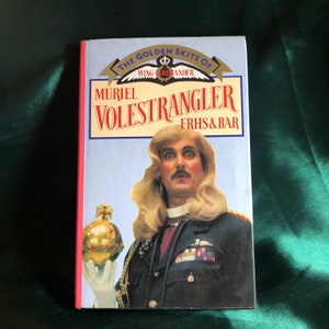 May include: A book cover with a blue background and the title "The Golden Skits of Muriel Volestrangler FRHS & BAR". The cover features a man with long blonde hair wearing a military uniform and holding a golden orb.