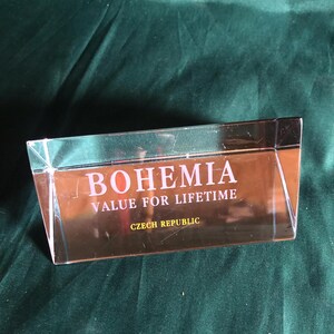 May include: A clear, rectangular glass object with the word "BOHEMIA" in white capital letters. Below, the text reads "VALUE FOR LIFETIME" and "CZECH REPUBLIC" in a gold colour. The object is on a dark green surface.