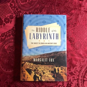 May include: A book titled "The Riddle of the Labyrinth" by Margalit Fox. The cover has a blue background with ancient symbols and the title in a vintage font. The bottom half shows a landscape with ruins. The book is a hardback.