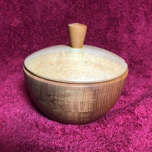 May include: A wooden bowl with a lid and a knob on top. The bowl is a light brown colour and the lid is a darker brown colour. The bowl is sitting on a red surface.