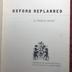 May include: A book titled "Oxford Replanned" by Thomas Sharp. The book cover features a black and white illustration of a coat of arms with the text "Fortis Est Veritas" and the words "Published for the Oxford City Council by the Architectural Press, London".