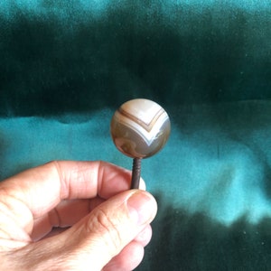 May include: A round, white agate knob with brown and white stripes, mounted on a metal pin.