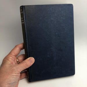 May include: A vintage hardcover book with a dark blue cover. The book's spine is black with white text. The book is held by a hand with a gold ring. The book's title is partially visible.