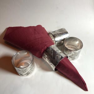 以下が含まれることがあります： バーガンディ色のナプキンと4つのシルバーのナプキンリングのクローズアップ。ナプキンリングには花の模様があります。ナプキンは折りたたまれ、リングに挿入されています。背景は白です。