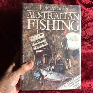 May include: A book titled "Jack Pollard's AUSTRALIAN FISHING" with fishing equipment on the cover. The cover shows fishing rods, tackle boxes, and a fish. The text "The classic bestseller now completely revised" is visible. The book is held by a person.