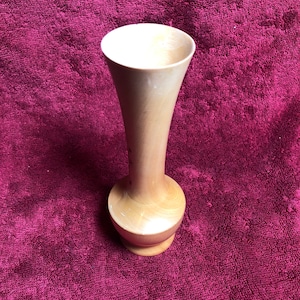 May include: A light brown wooden vase with a rounded base and a tall, slender neck. The vase is sitting on a dark red surface.