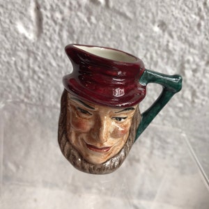 May include: A small ceramic pitcher with a whimsical face and a green handle. The pitcher is painted with brown, red, and white details.