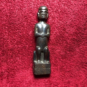 May include: A dark wood carving of a seated figure with a stylized face and hands. The figure is carved in a simple, geometric style and is sitting on a square base with carved details.