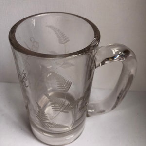 May include: A clear glass mug with a fern design etched into the side. The mug has a handle and a rounded bottom.