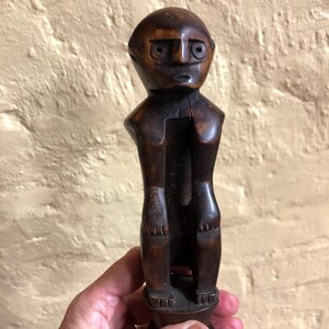 May include: A carved wooden figurine with a dark brown finish. The figure has a rounded head, stylized facial features, and a seated posture. The arms are bent, and the legs are together. The figurine is held in a hand.