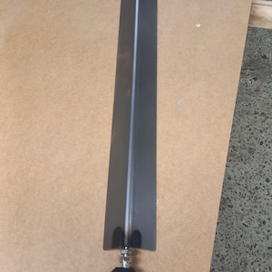 Op de afbeelding: Een lange, donkergrijze metalen kling met een punt en een zwarte rubberen voet. Het blad heeft een smalle, rechthoekige vorm met een verticale groef in het midden. Het lijkt een onderdeel van een grotere structuur te zijn.