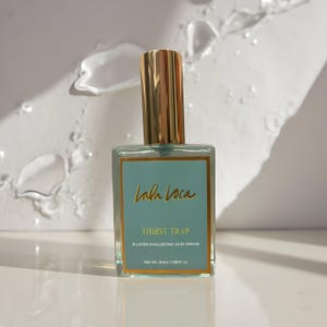 Op de afbeelding: Een rechthoekige glazen fles van Lala Loca Thirst Trap 8-laags hyaluronzuurserum. De fles heeft een gouden dop en een turquoise label met gouden letters. Op het label staat "Lala Loca", "Thirst Trap" en "8-LAYER HYALURONIC ACID SERUM". De fles bevat 50 ml.