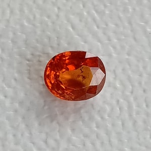 May include: Oval-cut, vibrant orange gemstone with multiple facets. The gemstone has a polished surface and reflects light, showcasing its brilliance. The gem is set against a white background, highlighting its color and clarity. This gemstone is suitable for jewelry.