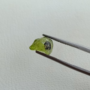 May include: A raw, light green peridot gemstone with a dark inclusion, held by silver tweezers. The rough-cut gem has a natural, irregular shape. The background is a plain, off-white surface. This image showcases a gemstone for jewelry making or collection.