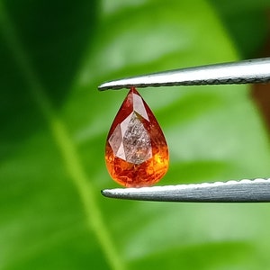 May include: A teardrop-shaped orange gemstone with a faceted cut, held by a pair of tweezers against a green background.