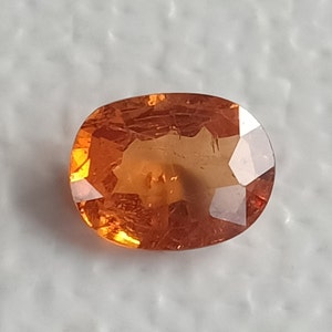 May include: A reddish-orange oval gemstone with a faceted cut. The gemstone is set against a white background.