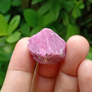 Puede incluir: Un cristal de rubí en bruto y sin pulir con un tono rojo rosado vibrante. La piedra presenta una estructura geométrica natural con facetas, estrías visibles y texturas cristalinas en toda su superficie, mostrando una apariencia opaca a translúcida.