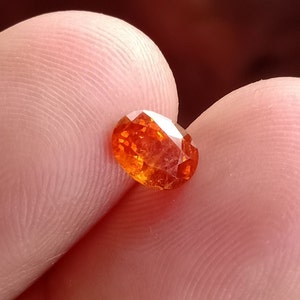 May include: A close-up of a bright orange oval-shaped gemstone. The gemstone is faceted and has a shiny, translucent appearance.