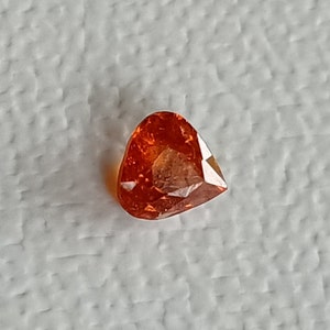 May include: A teardrop-shaped, faceted gemstone with a deep orange color. The gemstone is set against a white background.