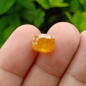 Puede incluir: Una piedra preciosa amarilla de corte cojín con una superficie facetada. La piedra muestra un tono naranja dorado vibrante y translúcido, sostenida entre dos dedos, destacando su corte geométrico pulido y sus reflejos naturales.