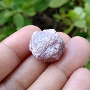 Puede incluir: Un cristal de rubí rosa con una forma geométrica facetada. El cristal es translúcido y tiene un ligero brillo.