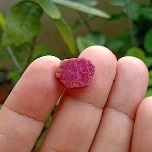 Op de afbeelding: Een kleine, ruwe, zeshoekige robijn met een diepe magenta-rode kleur en een getextureerd, mat oppervlak. De steen vertoont natuurlijke kristallijne facetten en een ondoorzichtig uiterlijk, wat de ongepolijste, ruwe minerale structuur benadrukt.