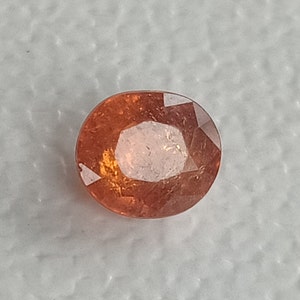 May include: A faceted, oval-shaped gemstone with a warm, orange-brown hue. The gem has a central, lighter-colored area and a polished surface, reflecting light. The gemstone is set against a neutral background.