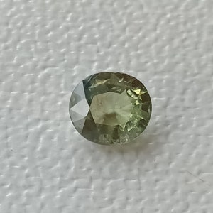 May include: A round, faceted green gemstone with a light green hue. The gemstone is set against a white background.