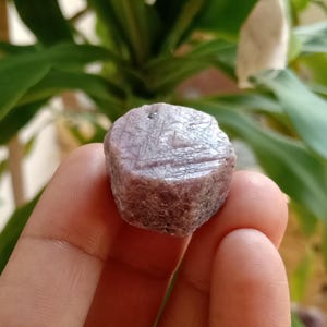 May include: A close-up of a purple, rough-cut gemstone with a hexagonal shape. The gemstone is held in a hand, and the background is blurred.