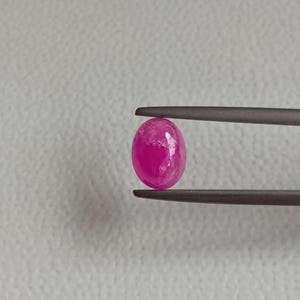 May include: A vibrant, oval-shaped, pink gemstone is held by tweezers against a neutral background. The gemstone has a polished surface with subtle internal inclusions, showcasing its natural beauty. This gemstone is ideal for jewelry making.