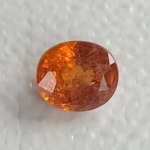 May include: An oval-shaped, orange gemstone with a reddish hue. The gemstone has a smooth, polished surface and is set against a white background.