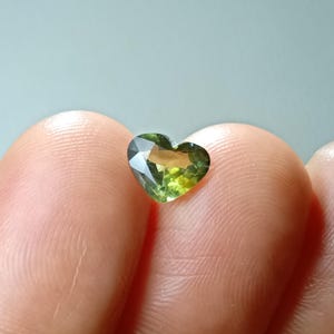 Pode incluir: Uma pedra preciosa em forma de coração com um corte facetado apresentando um gradiente de tons verde-oliva e amarelo-dourado. A pedra exibe reflexos de luz internos e clareza, com uma superfície polida de bordas nítidas e silhueta definida.