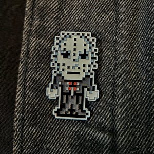 May include: Pixelated enamel pin depicting a character with a pale face, black suit, and red tie. The pin is attached to a dark denim jacket. The design is reminiscent of 8-bit video game graphics.