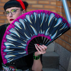May include: A large folding hand fan featuring a graphic print of sharp, white, and blue monster teeth against a black background with a vibrant magenta border. The fan has black ribs and is held open to display the detailed, jagged tooth pattern.
