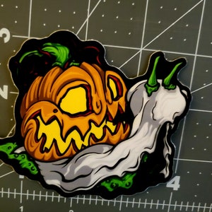 May include: A green and orange pumpkin jack-o'-lantern with a white ghost-like cloth draped over it. The pumpkin has a scary expression with a wide grin and glowing yellow eyes.