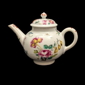 May include: White porcelain teapot with a lid, spout, and handle. The teapot is decorated with colourful floral patterns in pink, yellow, and green. The handle and spout have red accents. The teapot sits on a black background.