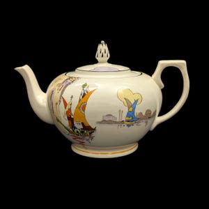 May include: A cream-colored ceramic teapot featuring a decorative illustration of colorful sailboats on water. The teapot has a curved spout, a sturdy handle, and a lid topped with a pointed, perforated finial. The design includes yellow, orange, and blue sails.