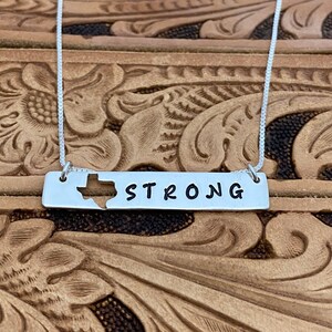 May include: A silver necklace with a rectangular pendant. The pendant has a cutout of the state of Texas and the word "STRONG" in black. The necklace is shown against a brown, textured background.
