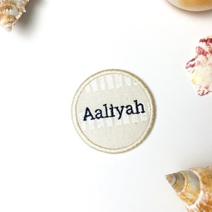 May include: A round, beige patch with the name "Aaliyah" embroidered in black. The patch has a striped pattern and a beige border. Seashells are arranged around the patch on a white background.