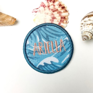 以下が含まれることがあります： オレンジ色で「ARIELLA」と刺繍された、青と白の丸いパッチ。パッチは波模様で、濃い青色の縁取りがあります。白い表面に、パッチの周りにいくつかの貝殻が配置されています。