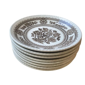 Puede incluir: Una pila de platos pequeños de cerámica blanca con un diseño floral marrón. Los platos tienen un borde decorativo y un motivo central detallado. Los platos están apilados ordenadamente, mostrando el intrincado patrón y el estilo clásico.