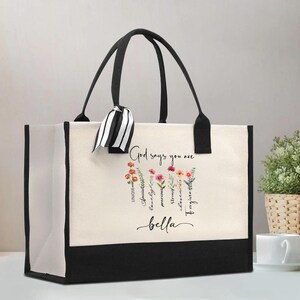 May include: A cream-coloured canvas tote bag with black trim and black handles. A black and white striped ribbon is tied to one handle. The front features a floral graphic with the text God says you are, followed by words like unique and bella.