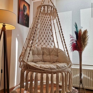 Puede incluir: Una silla colgante de macram&eacute; beige con un dise&ntilde;o circular, con asiento y respaldo tejidos. La silla tiene flecos en la parte inferior y est&aacute; suspendida de un anillo de metal. La silla est&aacute; en una habitaci&oacute;n con una l&aacute;mpara de pie y una pintura.