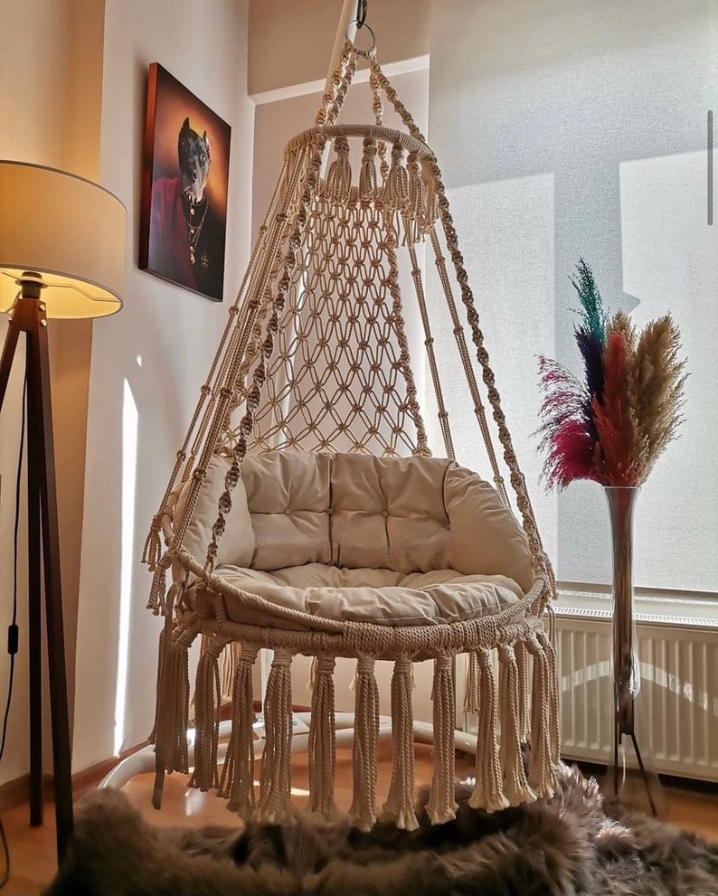 Puede incluir: Una silla colgante de macram&eacute; beige con un dise&ntilde;o circular, con asiento y respaldo tejidos. La silla tiene flecos en la parte inferior y est&aacute; suspendida de un anillo de metal. La silla est&aacute; en una habitaci&oacute;n con una l&aacute;mpara de pie y una pintura.
